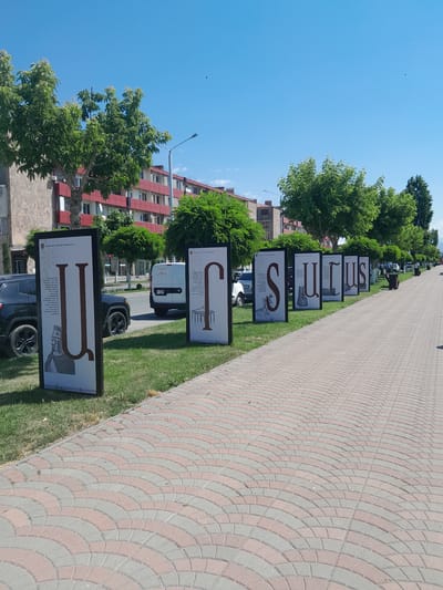 The image depicts a sunny day in Artashat, Armenia, showcasing an outdoor exhibition of large posters featuring Armenian alphabet letters and historical figures. These posters are lined up along a paved walkway, bordered by a grassy area with trees. The walkway is constructed of interlocking paving stones in a fan pattern, with alternating pink and grey colors. In the background, there are residential buildings with red and grey facades, and trees line the street. A white van and several cars are parked or driving on the road. The sky is a clear blue with a few distant birds. The exhibition appears to be set up for public viewing, likely a cultural or educational display in a public space within the city.
