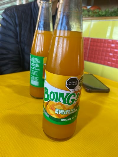 Two glass bottles of Boing! mango juice lie horizontally on a yellow surface. The bottle closest to the camera displays a bright yellow label with "Boing!" written in large, bold letters, accompanied by an image of mango slices. The label also includes nutritional information, specifically an "EXCESO CALORÍAS" warning, and states the juice contains 12% mango pulp. Text on the label reads "BEBIDA CON 12% DE PULPA DE MANGO" and "CONT. NET. 354 ml". A dark object is visible beside the bottles. A person wearing a black shirt is partially visible on the left. The background is a blur of green, yellow, and red.