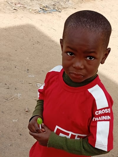 A young child with dark skin and a shaved head is depicted from the waist up, standing outdoors in Fanisau, Nigeria. The child wears a bright red short-sleeved t-shirt with white stripes on the shoulders and arms, layered over a green, long-sleeved shirt or turtleneck. The red t-shirt has white text on the right sleeve that reads "CROSS TRAININ", likely an abbreviation for "CROSS TRAINING". The child holds a small, round, green lollipop in both hands, positioned in front of their chest.
The child's expression is serious and direct, with large, dark eyes looking straight ahead at the viewer. There's a slight pout to the lips, conveying a contemplative or perhaps slightly somber mood.
The setting appears to be an unpaved outdoor area with light brown, dusty ground. In the background, there's some scattered debris and what looks like dried vegetation or waste materials, suggesting a less manicured environment. Natural daylight illuminates the scene, creating soft shadows, indicating it is daytime. The overall impression is one of a candid moment captured in a humble outdoor setting.