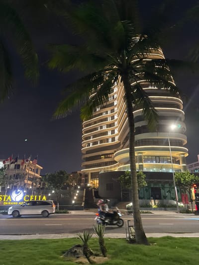 The image captures a nighttime street scene in An Hải Ward, Da Nang, Vietnam. A tall, modern hotel building, identified by its sign as "STA CELIA DA NANG HOTEL," dominates the background. The building features a curvilinear, multi-story design with horizontal bands of white LED lighting illuminating its exterior facade. Numerous windows are visible, some emitting light. In the mid-ground, a multi-lane asphalt road spans horizontally. A silver SUV is positioned on the left lane, and a motorbike with two blurred occupants, exhibiting motion blur, travels rightward in the closer lane. A prominent palm tree stands in the foreground, partially obscuring the hotel, with its fronds visible at the top. The immediate foreground features a grassy median strip with smaller, spiky plants and rocks. To the left of the main hotel building, a smaller structure displays multiple international flags, including those of Vietnam, Canada, Malaysia, Japan, and the United States. Artificial lighting from the building, streetlights, and vehicle headlamps illuminate the scene against a dark night sky; a green light artifact is visible in the upper left portion of the sky.