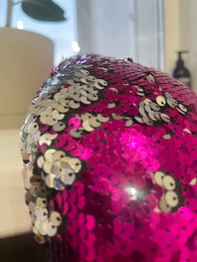This media file is a close-up shot of a round object covered in iridescent sequins. The sequins are predominantly pink and silver, creating a shimmering, reflective surface. The object appears to be curved, with the sequins arranged in an overlapping pattern. The background is blurred, but suggests an indoor setting with soft lighting. There is a hint of something dark and bottle-shaped in the upper left corner, out of focus. The lower part of the object rests on a textured, light-colored surface, possibly fabric or a smooth material. The overall impression is one of texture and vibrant color due to the reflective nature of the sequins. There is no discernible activity or event taking place; it is a static depiction of the object. No text is visible. The location context provided is Gorodets, Russia, but this information is not visually evident from the image itself.