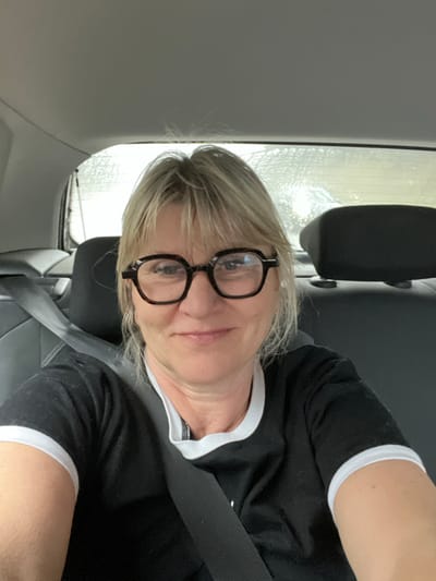 The image displays an adult female in the rear passenger compartment of a vehicle, located in Saint-Maximin-la-Sainte-Baume, France. The woman, positioned centrally, has blonde hair with bangs and wears octagonal tortoiseshell-patterned eyeglasses. She is depicted from the chest up, looking directly towards the camera with a slight, closed-lip smile. She wears a black short-sleeved t-shirt with white piping visible on the collar and sleeves. A black seatbelt is secured across her left shoulder and chest. The vehicle's interior includes dark gray seating and headrests, with a light gray ceiling panel. The rear window, visible behind the woman, shows a blurred, light-colored external environment and appears to have moisture or smudges on its surface.