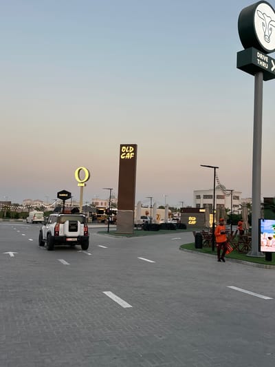 The image depicts an outdoor scene at dusk, likely in Ras Al Khaimah, United Arab Emirates. A street with brick paving and white directional arrows leads the viewer through a bustling area with several small eateries or food stalls. The sky is a soft gradient of pale blue and beige, indicating the transition from day to night.
Prominently displayed are various signs. A "DRIVE THRU" sign with a cow graphic stands tall on a pole. Further along, a tall, dark sign with illuminated yellow text reads "OLD CAF". Another distinctive sign features a large, illuminated yellow ring above a smaller sign that says "SADDLE".
Several white vehicles are parked or driving on the street, suggesting a popular destination. In the background, there are buildings with Mediterranean-style architecture, and outdoor seating areas with umbrellas and chairs. A few people are visible, some in bright orange vests, possibly staff members or patrons. One person is sitting at a table, and another appears to be walking away from the camera.
The overall atmosphere seems relaxed and social, characteristic of an evening gathering place. The lighting from the signs and some string lights creates a warm and inviting ambiance. The presence of multiple food vendors and the drive-thru suggests a place catering to casual dining and quick service.