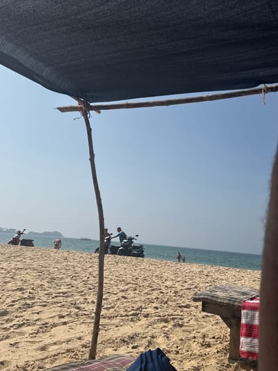 A beach view through a makeshift shaded structure. A dark material is attached to a wooden frame. The beach has light-colored sand. Motorbikes are parked near the shore. People are visible walking and standing by the water. A cloth-covered bench is partially visible in the foreground. The sky is clear and light blue. The image is taken from a slightly elevated perspective.