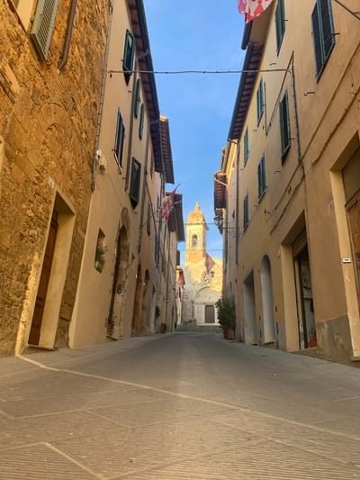 The image captures a narrow street in the Italian town of San Quirico d'Orcia, Tuscany. The perspective is low to the ground, looking up the cobblestone street towards a church at the end. Buildings line both sides of the street, their facades painted in warm, earthy tones of yellow and ochre. The building on the left is constructed from rough, honey-colored stone, while the one on the right is stuccoed. Both feature traditional green shutters on the windows. Red and white checkered flags are displayed from some of the buildings. The street is empty of people, bathed in the warm light of what appears to be late afternoon or early morning sun, casting long shadows across the cobblestones. The sky above is a clear, bright blue. The church at the end of the street has a bell tower and an arched entrance, with steps leading up to it.