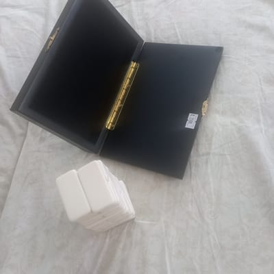 An open black rectangular box with gold hinges sits on a white fabric surface with a floral pattern. The box is open at a roughly 45-degree angle, with the left side elevated and the right side flat. Inside the left side of the box appears to be a dark, possibly velvet lining. The right side of the box is also lined with black material and has a small white sticker with "PYB 1500" written on it in black text, placed near the right edge. To the left of the box, a stack of approximately ten white rectangular objects, resembling dominoes or game tiles, is arranged in a staggered fashion. The background is a textured white fabric, possibly a tablecloth or bedding, with a subtle, light grey floral or swirling pattern. The lighting is soft and diffused, suggesting an indoor setting. The overall impression is of a product display or a neatly arranged collection of items. The image does not provide any information or context related to Cepeleuți, Moldova, or any specific location.