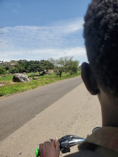 The image shows a person's head and shoulder in the foreground, looking forward. The person is riding a motorcycle on a paved road. The road curves to the left and is bordered by a grassy area with rocks and trees on the right. In the background, there are more trees and rocky hills. The sky is blue with some white clouds. The scene appears to be in a rural or semi-rural area. The overall impression is one of travel and observation of the surrounding landscape.