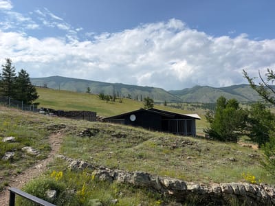 The image depicts a rustic scene in the village of Sarma, Russia. In the foreground, a rocky, grassy hillside is dotted with small yellow wildflowers. A dirt path winds its way up the hill. In the midground, a dark, modern-looking house with a white satellite dish is situated on a grassy slope. The house has a covered porch with vertical gray slats. Beyond the house, a wooden fence encloses a fenced area with some stacked wooden materials. Rolling green hills and mountains, some covered in dark forests, form the backdrop under a partly cloudy sky. Several evergreen trees are scattered across the landscape.