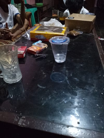 The image shows a dimly lit table at night, likely indoors given the lack of any outdoor environmental cues. On the table, there are several objects, including a glass mug, two packs of cigarettes, a box labeled "SPECIAL BUTTER," a plastic cup with a blue lid, and a small plastic bag containing some food item. There is also a cardboard box in the background. The table surface is dark and appears to be somewhat dirty or smudged.

In the background, a person wearing a white t-shirt with blue trim and a Spotify logo is visible. Their hands are resting on their lap, and their posture suggests they are sitting. Another person's head is visible in the far background, also appearing to be seated. The overall impression is a casual gathering or moment of relaxation.

The time of day is indicated as night by the darkness. There are no clues about the weather. The presence of cigarettes and a beverage in a cup suggests a moment of leisure or conversation. The "SPECIAL BUTTER" packaging and the small bag of food might indicate snacks or a shared meal. The scene feels informal and domestic, possibly in a home or a casual eatery. The lighting is low, creating a sense of intimacy or quietude. The mood appears relaxed, with no obvious signs of stress or urgency.