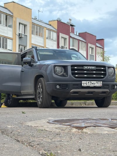 A dark gray Haval SUV is parked on a paved road in front of a row of colorful buildings. The driver's side door of the SUV is open. A person wearing yellow shoes and dark pants is partially visible standing next to the open door. The SUV has a license plate that reads "y 417MY 9977 RUS". The buildings in the background are multi-story with yellow, red, and beige facades, featuring numerous windows and visible air conditioning units. The sky is overcast with clouds. A manhole cover is visible on the road in the foreground. The overall scene appears to be in a residential urban environment.