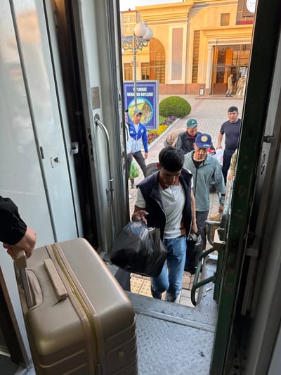 The image, taken from inside a train, shows a group of people on the train platform in Jizzakh, Uzbekistan. A person, presumably a passenger, is pulling a beige suitcase with wheels and an extendable handle into the train. The person is also holding a black plastic bag. Outside the train, several people are standing, some appearing to be waiting to board or disembark. One man is wearing a black t-shirt. Another man is wearing a blue baseball cap. A third man wears a blue baseball cap. A fourth man is wearing a blue athletic jacket. A nearby building has intricate architectural details, including arches and latticework. A poster is visible. Streetlights with ornate bases are positioned along the sidewalk.