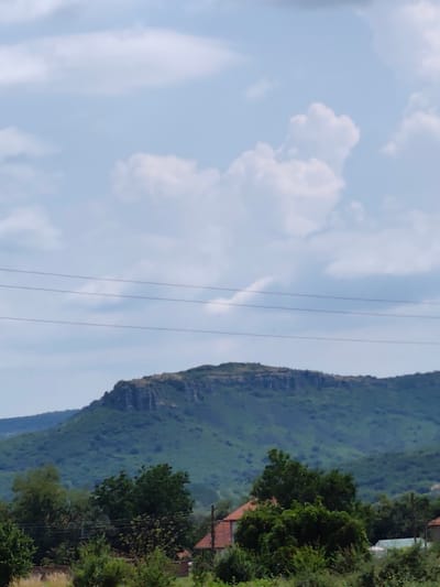 A landscape photograph captures a view of a rocky plateau mountain, known as the "Krumovitsa rock formations," located near the village of Umarevtsi, Bulgaria. The mountain's summit features a distinct, flat, and eroded rock face. The lower slopes are covered in lush green vegetation, with trees and shrubs creating a dense forest. In the foreground, a few residential buildings with terracotta roofs are visible amidst the foliage, suggesting a rural setting. Overhead, a cloudy sky with scattered white cumulus clouds completes the scene. Power lines are strung across the sky, indicating the presence of infrastructure in the area. The overall impression is of a serene natural landscape with hints of human habitation.