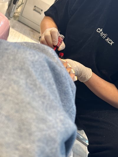 A person wearing a black t-shirt with "charli xcx" printed on it is applying red nail polish to a person's toenails. The nail technician is wearing disposable white gloves. The recipient of the pedicure is wearing denim pants. The scene appears to be indoors. It is not possible to determine if the location is in Strömstad, Sweden based on the provided information.