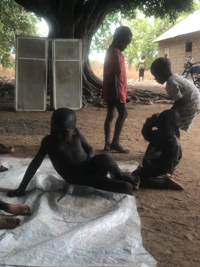 The image depicts an outdoor scene in Ninzo South, Nigeria, featuring multiple individuals and objects under the shade of a large tree.
**Environment:** The ground is composed of reddish-brown dirt. A large tree with extensive, visible surface roots dominates the left and central background, providing significant overhead foliage. In the far background, a single-story building constructed of light brown, unpainted brick or mud is visible on the right. Sparse, dry vegetation and what appears to be a field or open area are visible beyond the tree and building. The lighting suggests an overcast sky or heavy shade.
**Objects:**
* Two large, rectangular speaker boxes, light gray with perforated grilles, stand upright in the mid-ground to the left of the tree.
* A white, textured tarp or sheet is laid on the ground in the immediate foreground, extending from left to center.
* A pair of light brown slip-on sandals or slippers are on the dirt ground to the left of the tarp.
* A blue motorcycle is partially visible, parked to the right of the building in the background.
**People:** There are five children and one adult visible.
* **Child 1 (Foreground Left):** A young, dark-skinned child is lying on their left side on the white tarp, facing slightly away from the camera. They are shirtless and wearing dark shorts, with their legs bent.
* **Child 2 (Foreground Right):** A young, dark-skinned child is sitting on the dirt ground next to the tarp, wearing a dark long-sleeved shirt with a white Adidas logo and what appear to be red or pink shorts. Their hands are raised towards their head.
* **Child 3 (Mid-ground Left):** A barefoot, dark-skinned child stands upright, wearing a red t-shirt and dark shorts, looking towards the right side of the frame.
* **Child 4 (Mid-ground Right):** A barefoot, dark-skinned child stands, bent forward, wearing a light-colored patterned t-shirt and shorts. They are looking down at Child 2.
* **Child 5 (Background Right):** A small, dark-skinned child is standing further back, near the building, partially obscured by the tree roots.
* **Adult (Background Far Right):** An adult figure is standing in the distance near the building, wearing a horizontally striped top.
**Actions/Interactions:** Child 1 is resting on the tarp. Child 2 is sitting on the ground with hands near their head. Child 4 is bending over, seemingly interacting with Child 2. Child 3 and Child 5 are standing passively. The adult in the background is standing at a distance.