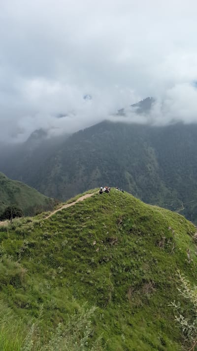 A group of people are gathered on a grassy hilltop in the mountains of Cheelaage, India. The sky is overcast with clouds obscuring the peaks of the distant mountains. The hilltop is covered in lush green grass, and a dirt path winds around its crest. The people are sitting or standing, appearing to be enjoying the view. Some are dressed in casual clothing, and one person appears to be wearing a white shirt and dark pants. The overall scene is one of serene natural beauty, with the overcast sky adding a sense of drama to the mountainous landscape.