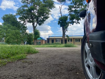 An outdoor, low-angle, eye-level shot captures a rustic scene on a dirt road in Tagarnogsky, Russia. In the foreground, a portion of a dark red car is visible on the right, with its silver wheel and part of the bumper in focus. The car is positioned on a dirt and gravel path that extends into the background. To the left of the road, a dense area of green grass and weeds grows, with some dry twigs and leaves scattered on the path.
In the mid-ground, two trees with full green canopies stand tall, their branches reaching towards the sky. Behind the trees, a long, low building with a yellow facade and a light blue roof is visible. The building has several windows with dark frames and what appear to be closed doors. A small brick structure with a blue roof is situated to the left of the yellow building. A wooden fence runs along the edge of the property.
The background features a bright blue sky dotted with fluffy white clouds, suggesting fair weather. Power lines are visible crisscrossing the sky between the trees. The overall impression is one of a rural or suburban setting in Russia, possibly a school or administrative building in a less developed area. There are no people visible in the image.