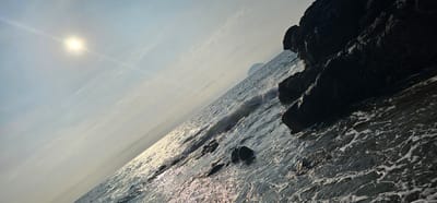 The image, taken from the coast of Girvan, United Kingdom, depicts a sunlit ocean with rocky outcrops and a distant island. The sun, a bright disc with a distinct lens flare, is positioned in the upper left quadrant, casting a shimmering path across the water. The sea itself is choppy, with waves breaking against dark, angular rocks that dominate the right side of the frame. In the distance, a small, rounded island is visible on the horizon, partially obscured by the haze. The sky above is a pale blue, streaked with wispy clouds. The overall impression is one of a rugged coastal landscape bathed in the glow of the sun.