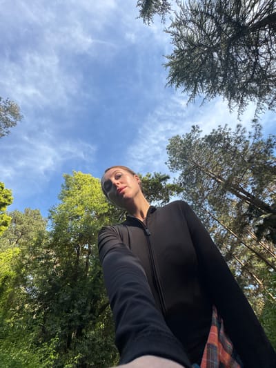 An upward-angled outdoor photograph shows a person wearing a black long-sleeved garment with a glimpse of a plaid pattern visible near the right edge. The person's face is in the center-left, with a blurred-out area obscuring one eye. The background consists of a blue sky with thin white clouds, framed by green trees, some appearing sparse with branches, and others appearing dense with foliage. The location is unidentifiable as Tsanbaikwara, Abkhazia, with the provided image.