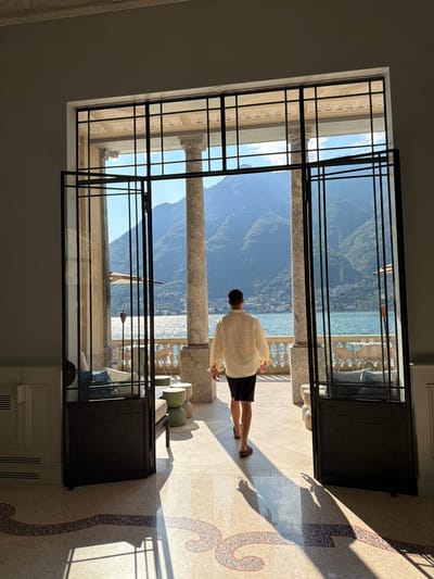 A person walks out of a building through an open doorway. The person is wearing a cream-colored long-sleeved shirt and black shorts, and appears to be walking towards a balcony overlooking a body of water and mountains. The doorway is framed by large columns and an ornate metal structure, and the floor inside the building is tiled with a mosaic pattern. The balcony outside has tables and chairs, suggesting a cafe or restaurant setting. The background shows a bright, sunny day with a clear blue sky over the water and lush green mountains. The scene appears to be in Pognana Lario, Italy, likely at a villa or hotel with a view of Lake Como.