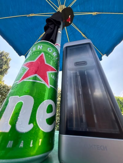 The image shows a close-up, low-angle shot of a Heineken beer can, a black baseball cap with a red bull logo, and a portion of a blue umbrella, all positioned under a clear sky with some greenery in the background. A sleek, rectangular device with a transparent cover and the text "QUXTECH" is also visible. The Heineken can is a prominent subject, with its iconic green label, red star, and condensation suggesting it is cold. The can is tilted, with the brand name "HEINEKEN" and "ORIGINAL" clearly legible. The black baseball cap has a red and white embroidered logo of a bull's head. The blue umbrella, with its yellow ribs, provides shade. The QUXTECH device appears to be some sort of electronic equipment, possibly a scanner or a camera. The scene is outdoors, likely on a sunny day, given the bright sky and the condensation on the can. The presence of the umbrella and the beer can suggests a relaxed, casual outdoor setting, possibly a park, a patio, or a garden, during daytime. No people are visible in the frame. The overall mood is relaxed and summery. The location context is Vicenza, Italy, but there are no specific visual cues in the image to confirm this.