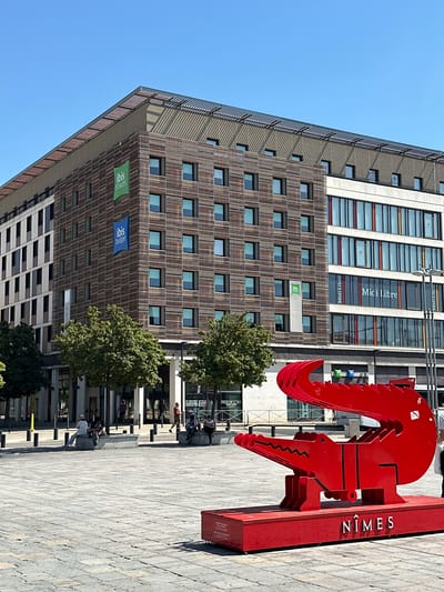 The image shows a bright, sunny day in Nîmes, France. In the foreground, a large, vibrant red metal sculpture of a crocodile stands on a red platform. The word "NÎMES" is prominently displayed in white lettering on the front of the platform. Behind the sculpture, a modern building complex rises, featuring a facade of horizontal wooden slats on one section and large glass windows with colorful accents on another. Several Ibis hotel signs are visible on the building. Trees line the street in front of the building, and people are seen sitting on stone benches in the plaza. The ground is paved with large stone tiles. The sky is a clear, brilliant blue.