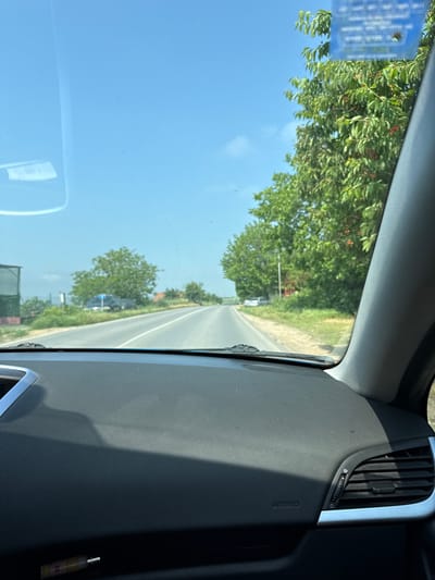 The image depicts a bright, sunny day seen from the interior of a car, looking forward through the windshield. The perspective is from the front passenger side.
In the foreground, the car's dark gray dashboard is visible on the right side of the frame, featuring a textured surface. An air vent with silver trim is prominently displayed on the upper right, accompanied by a small dial with a power symbol. The word "AIRBAG" is embossed on the dashboard panel below the vent. In the lower right corner, a small, decorative bottle, possibly containing perfume or an air freshener, sits on the dashboard.
Through the windshield, a straight, paved road extends into the distance, flanked by abundant green trees and foliage on both sides. The trees are dense, with some displaying small red berries among their leaves. The sky above is clear and a vibrant blue, indicating fair weather. A rectangular blue sticker with white, illegible text is affixed to the upper left corner of the windshield. Light reflections are visible on the windshield, particularly in the lower left area.
The scene suggests a drive through a rural or semi-rural area near Ezerovo, Bulgaria, under bright mid-day or early afternoon sunlight, given the strong illumination and clear sky. No people are visible, and the activity is implied to be traveling by car.