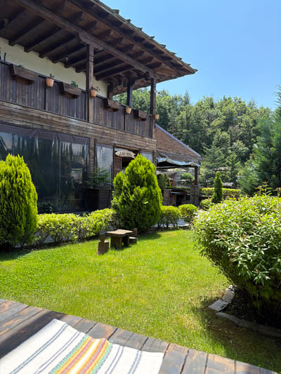 This image captures a serene outdoor scene, likely a garden or courtyard belonging to a traditional establishment in Leshten, Bulgaria. The setting is dominated by a rustic wooden building on the left, featuring dark timber planks and an intricately designed sloping roof with visible wooden beams and supports. Attached to the building are several wooden planters, some holding terracotta pots with small red flowers. Large windows on the ground level of the building are covered with transparent, possibly plastic or tarp, material, suggesting an enclosed seating or dining area. A wooden sign with Cyrillic text "ДЯКОВЪ" (Dyakov) hangs above one of these windows.
To the right of the building, a meticulously maintained, vibrant green lawn stretches across the middle ground. The lawn is bordered by various types of lush green bushes, some neatly trimmed into hedges, while others appear more natural. Further in the background and along the edges of the scene are numerous tall, mature trees, indicating a forested or heavily vegetated environment beyond the immediate garden. Several conical evergreen shrubs are also integrated into the landscaping, and a small, rustic wooden bench is visible on the lawn. A low stone wall or border can be seen outlining a section of the lawn next to some bushes.
In the immediate foreground on the right, a dark wooden surface, possibly a table or railing, is visible. Placed on this surface is a folded white cloth adorned with horizontal stripes in various colors including blue, green, yellow, orange, and red.
The scene is bathed in bright, clear sunlight, casting distinct shadows and suggesting a beautiful, sunny day, likely during late morning or early afternoon. There are no people visible, contributing to the tranquil and peaceful ambiance. The overall impression is one of natural beauty and traditional architecture, characteristic of a picturesque Bulgarian village.