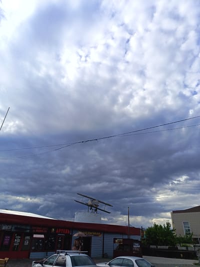 A low-angle, full-shot photograph captures a dramatic sky filled with dark, heavy clouds, hinting at an approaching storm. Below the turbulent sky, a low-rise commercial building with a red roof is visible. The building features a prominent sign with "АРТЕЛЬ" in large, bold letters, and other text that includes "МЯСО" and "ТОРГОВЫЙ ЦЕНТР ул. ЛЕНИНА 114". To the right of "АРТЕЛЬ", a smaller sign reads "ШАШЛЫЧНАЯ Улётная". Mounted atop a brick structure on the roof of the building is a vintage biplane model, adding a quirky detail to the scene. In front of the building, a silver sedan is parked. The overall atmosphere is overcast and moody, likely indicating late afternoon or early evening given the lighting conditions. The presence of the biplane and the commercial establishments suggests this is a roadside strip or a small business complex in Slavgorod, Russia, possibly on a street named Lenina. The image evokes a sense of anticipation for the weather to change.