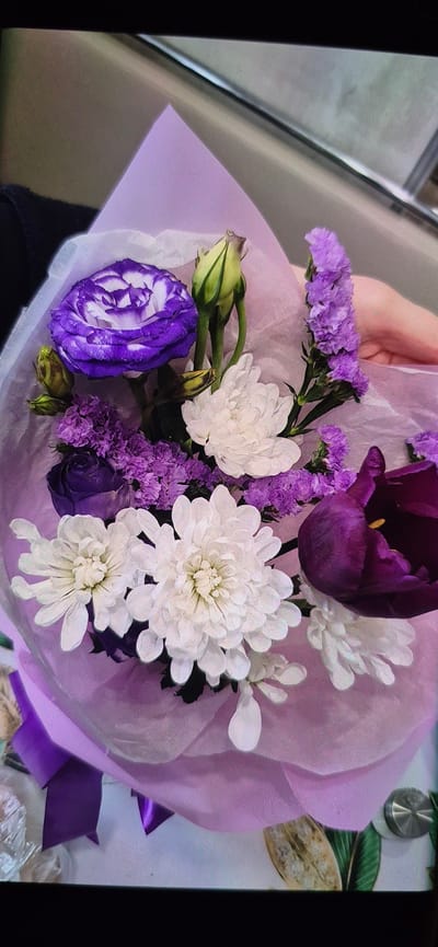 This is a close-up, top-down shot of a bouquet of flowers, primarily in shades of purple and white, wrapped in delicate purple and white paper. The bouquet is held by a person's hand, partially visible on the right side of the frame. The flowers include a large purple and white rose, several white chrysanthemum-like flowers, a dark purple tulip, and smaller sprigs of purple flowers. The bouquet is presented against a muted, neutral background that appears to be an indoor setting with some reflective surfaces and a hint of greenery and decorative elements at the bottom edge. There is no discernible information about the location being Yemelyanovo, Russia, nor are there any details about the people or actions depicted beyond the presentation of the bouquet.