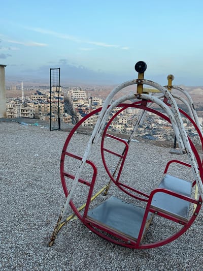 An old, rusty merry-go-round with two seats sits on a gravel surface. The structure is red and white, showing signs of wear. In the background is a cityscape with buildings clustered together, extending into the distance towards a horizon of hills under a pale sky. A rectangular metal grid is partially visible in the lower left, adding another element to the scene's composition.