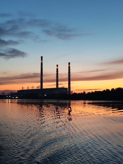 The image shows a twilight scene at the Konakovo State District Power Plant in Konakovo, Russia. Three prominent smokestacks stand silhouetted against a gradient sky of blues, grays, and oranges, with the sun setting on the horizon. The smokestacks are illuminated from within. A dark industrial building complex stretches across the middle ground, also silhouetted. In the foreground, a body of water reflects the colors of the sky and the industrial structures, with ripples disturbing the surface. Trees form a dark, silhouetted line along the far bank of the water. There are no people or animals visible in the scene. The overall atmosphere is one of industrial twilight.