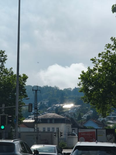 The image depicts a street scene in Lörrach, Germany, on a sunny day with clouds. In the foreground, a traffic light with a green signal is visible, along with two cars. To the left, a tall pole stands prominently. Behind these, a hilly landscape is covered with houses and trees, with the sun reflecting brightly off some rooftops. Several electrical poles and wires are scattered throughout the scene. A billboard advertising "Rossmann" is partially visible on the right side, displaying text in German. The overall impression is that of a suburban or city fringe area during daytime.