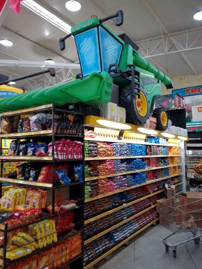 The image shows the interior of a supermarket. The most prominent feature is a large, green, toy-like combine harvester replica displayed above a shelf stocked with various snack bags. The harvester has a blue tinted windshield and large yellow wheels with black tires. Below the harvester, shelves are packed with colorful snack bags, including chips and cookies, arranged neatly. In the foreground, shopping carts and cardboard boxes are visible. The supermarket appears to be well-lit with overhead fluorescent lights. The environment is that of a typical retail store. There are no people visible in the image. Based on the sign visible in the background which reads "MERCADO SÃO GABRIEL," this supermarket is likely located in São Gabriel do Oeste, Brazil.