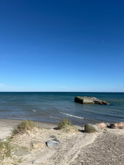 The image shows a beach landscape, potentially near Skagen, Denmark, given the coastal environment. The scene is vertically oriented. The left side features a clear, bright blue sky, with a crescent moon faintly visible. Transitioning to the right, the sky meets a body of water, likely the sea. The water shows gentle wave activity and a light to medium blue hue. A concrete structure, possibly a breakwater or remnant of coastal defense, stands partially submerged in the water. The right side of the image displays the shoreline, a mix of sand, small rocks, and coarse vegetation. The ground transitions from sandy beach to a gravelly, more solid terrain near the top right corner. No people are visible in the image.