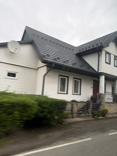 The image shows a white, two-story house with a dark gray tiled roof, located on a cloudy day in Wilkowisko, Poland. A satellite dish is mounted on the left side of the house, near the corner. The house has horizontal white siding and several windows with dark frames. A red front door is visible, as well as a staircase leading up to it, with a black metal railing. A green hedge runs along the front of the property, partially obscuring the lower level of the house and the surrounding area. The foreground shows a wet asphalt road with a white line, suggesting it may have recently rained. The overall scene depicts a residential exterior in what appears to be a suburban or rural setting.