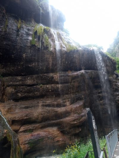 The image captures a majestic waterfall cascading down a colossal rock face in the city of Khushto-Syrt, Russia. The rock formation is characterized by its dark, damp texture, accentuated by streaks of green moss clinging to its surface. Layers of sedimentary rock are clearly visible, creating a rugged and textured appearance. Water streams down from the top, creating a misty veil that partially obscures the upper reaches of the cliff. At the bottom of the image, a metal railing and a pathway indicate a man-made structure, likely a viewing platform or bridge, designed to allow visitors to safely approach the waterfall. Lush green vegetation and a scattering of small yellow flowers are visible near the base of the rock face, adding a touch of natural beauty to the scene. A dark, rectangular signpost stands near the railing, its purpose unclear from this perspective. The sky above is overcast and bright, suggesting a cloudy or misty day.