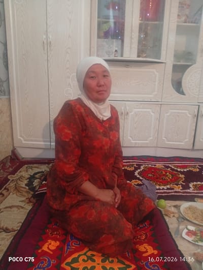 A woman wearing a white hijab and a red floral patterned dress is sitting on a rug in a room. Behind her, a light-colored wooden cabinet with glass doors is visible. The rug is dark red with a complex pattern. A plate with food and a small green apple are on the floor to the woman's right. The timestamp "16.07.2026 14:36" is visible in the lower right corner of the image, suggesting the photo was taken on this date and time. The overall setting appears to be indoors, possibly a living room or dining area, in Чоң-Гара, Kyrgyzstan.