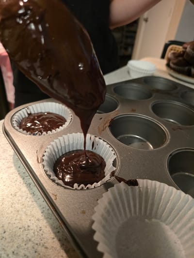 In this close-up, overhead shot, a dark, glossy liquid, likely melted chocolate, is being poured from a flexible container into white cupcake liners that are set inside a muffin tin. The muffin tin, made of a metallic material, has several circular indentations, two of which are occupied by the cupcake liners, each containing a portion of the chocolate filling. The chocolate is smooth and reflects the light, suggesting it is freshly melted. The scene appears to be in a kitchen environment, as indicated by the speckled countertop and a hint of cabinetry in the background. A person's arm, mostly obscured and wearing dark clothing, is visible on the left side of the frame, holding the pouring container. The lighting suggests it could be daytime, but there are no direct cues like windows or natural light sources. The overall impression is a candid snapshot of baking in progress. There is no visible text in the image.