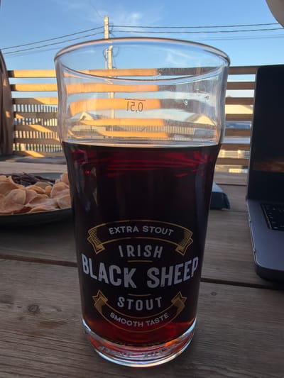 A close-up, eye-level shot shows a dark Irish stout in a clear glass on a wooden table. The glass has "EXTRA STOUT IRISH BLACK SHEEP SMOOTH TASTE" printed on it in yellow and white lettering. The stout is a deep reddish-brown color. In the background, a wooden fence and a blue sky with some wires are visible. To the right of the glass, a portion of a laptop is visible. To the left of the glass, a plate of light-colored chips and dark food is visible. The scene appears to be outdoors, possibly on a deck or patio. There is no indication of the specific city or location, except that it's in Russia.