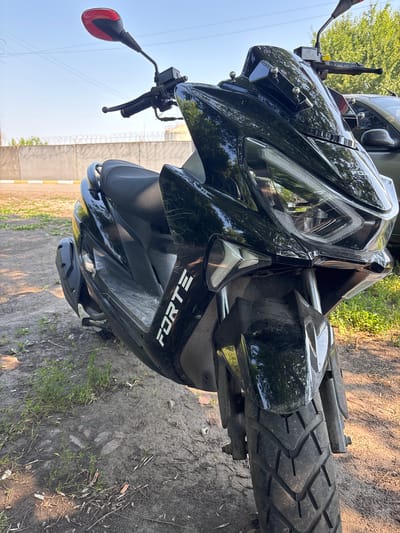 A black scooter is parked on a dirt ground, with grass and debris visible around it. The scooter has a red-tipped rear-view mirror, a black seat, and a prominent "FORTE" logo on its side. In the background, there is a concrete wall topped with barbed wire, suggesting a secured or industrial area. Beyond the wall, trees and a clear sky are visible. A portion of another vehicle, possibly a car, is parked to the right of the scooter. The scene appears to be outdoors, in daylight, and has a somewhat rural or unkempt urban feel. The location is identified as Pereiaslavske, Ukraine.