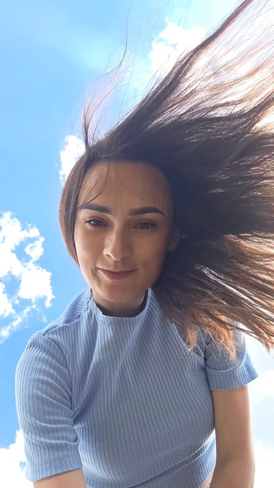 The media file is a close-up, upside-down selfie of a young woman with long, dark brown hair against a bright blue sky with scattered white clouds. She is wearing a light blue, ribbed, short-sleeved top. Her face is tilted upwards towards the camera, and she has a slight smile. Her eyes are brown, and she is wearing subtle makeup. Her hair is spread out, framing her face and suggesting movement, possibly from wind. The image appears to have been taken outdoors on a sunny day, likely during daylight hours. There are no visible text elements in the image. The setting appears to be an open outdoor space, with the sky being the dominant background element, indicative of a clear, pleasant day. The overall mood is relaxed and perhaps joyful.