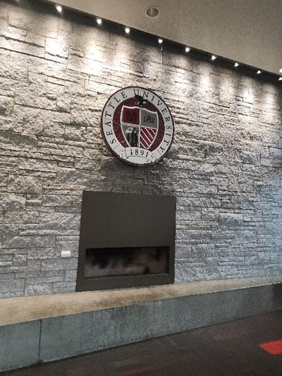 The image displays an interior scene featuring a prominent wall constructed from stacked, irregular grey stone veneer. Centered on the upper half of this wall is a large, circular emblem with "SEATTLE UNIVERSITY 1891" inscribed around its perimeter, enclosing a crest that includes a castle, the IHS Christogram, and other heraldic elements. Directly below the emblem, a rectangular, dark-colored modern fireplace is set into the stone wall; the firebox appears dark and empty. A white electrical outlet is visible on the stone wall to the left of the fireplace. Along the base of the wall, a long, low, grey concrete bench or planter extends across the frame. The ceiling above the wall is light-colored and features both recessed circular downlights and a linear arrangement of small, surface-mounted LED lights that illuminate the stone and emblem. The floor in the foreground is dark, with a faint red rectangular marking visible towards the bottom right. This interior is located within a building associated with Seattle University, in Seattle, Washington, United States. There are no people present in the scene.