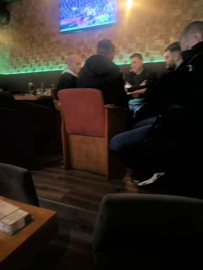 A group of people is gathered indoors, possibly in a bar, watching a television screen that displays a vibrant scene with green and blue hues. The room has a tiled wall illuminated with green light. A man in a suit sits in a maroon chair. A wooden table is visible on the floor. Dark sofas and armchairs suggest a comfortable, lounge-like atmosphere. Some people are standing, with one individual wearing blue jeans and white sneakers.