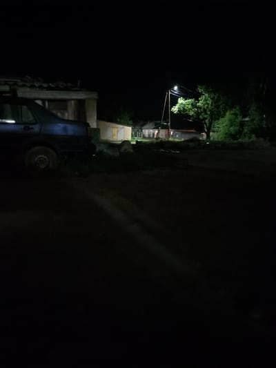 This is a nighttime photograph taken in Burma-Suu, Kyrgyzstan. The scene depicts a dimly lit rural area with a few buildings and a car. The foreground is dark, with shadows cast across a dirt road. On the left, the rear of a dark-colored car is visible, parked next to a dilapidated structure with a corrugated roof. Further into the scene, a streetlamp illuminates a large, leafy tree. Behind the tree, several houses are visible, appearing as simple, possibly residential structures. The overall atmosphere is quiet and still, suggesting a peaceful rural evening. There is no visible text in the image.