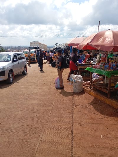 A bustling roadside market scene under a cloudy sky. A silver car and yellow taxi sit adjacent to a road lined with people, some standing near a large white truck. Market stalls with red umbrellas display various goods. A person with a backpack stands near bags, seemingly waiting. Another person in a red skirt is interacting with a vendor at a stall, filled with an assortment of items.