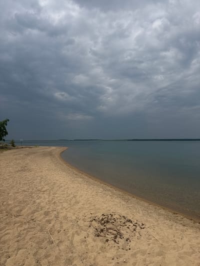 The image depicts a serene, natural landscape with a distinct division between sky and water. On the left side, a dramatic, overcast sky dominates the frame, filled with heavy, grey clouds that suggest impending rain or a recent storm. The light is diffused and muted, creating a somber atmosphere.

The right side of the image features a tranquil body of water, likely a lake or sea, meeting a sandy shoreline. The water is calm and reflects the muted tones of the sky, appearing a soft blue-grey. The shoreline is composed of light-colored sand, with a few scattered pieces of dry grass and debris, including a small, circular patch of dark, charred material that hints at a past campfire. The transition between the water and the sand is smooth and gradual, creating a sense of peaceful coexistence between the elements.

In the distance, a thin line of greenery marks the far shore, adding depth to the scene. A small tree with dark green foliage is visible near the bottom left corner, its branches reaching upwards as if bracing against the weather. The overall impression is one of natural beauty and quiet solitude. The time of day is difficult to ascertain precisely due to the overcast conditions, but it appears to be daytime. The weather is clearly cloudy and possibly humid, contributing to the subdued mood of the image.

No people or specific objects are prominently featured, allowing the viewer to focus on the natural elements. The setting suggests a remote or natural location, possibly a beach or lakefront. The emotional tone is calm and contemplative, perhaps a bit melancholic due to the heavy clouds. There is no visible text in the image.