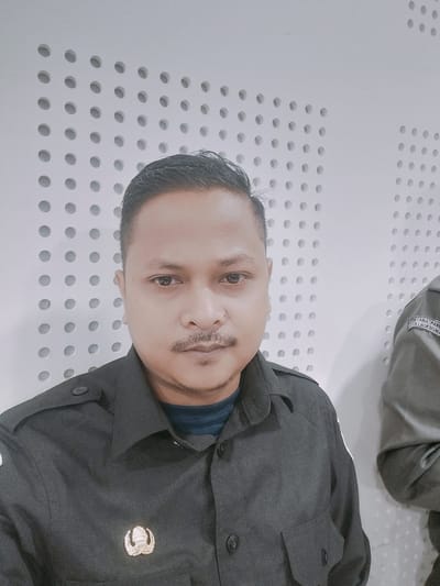 The image is a medium shot selfie of a man in Reuleut Timu, Indonesia. The man is wearing a dark grey uniform shirt with a gold emblem on the left chest. The background consists of a white wall with a pattern of evenly spaced, circular indentations. To the right of the man, another person in a similar grey uniform is partially visible. The lighting is bright, casting minimal shadows.