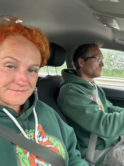 In the interior of a car, a woman with bright orange hair and blue eyes is in the foreground, looking at the camera and smiling. She is wearing a green hoodie with a white drawstring and a design on the front. A seatbelt is visible across her chest. To her right, a man wearing glasses and a similar green hoodie is driving. He is looking forward, out of the car. The car appears to be on a road, with trees visible in the background. The overall scene suggests a couple traveling by car. The location is identified as Tatarstanskie Chelny, Russia.