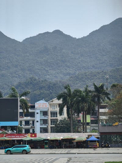 The image shows a street scene in Thành phố Hà Giang, Vietnam. In the foreground, there is a teal-colored SUV parked on the side of the road, next to stalls set up under colorful awnings. These stalls appear to be street vendors. Behind the stalls, there are several buildings, including one with a sign advertising "TAXI HA GIANG" and a phone number. Palm trees are interspersed among the buildings. A sign reads "XUÂN BÍNH NGỌ 2026". In the background, a large mountain covered in dense vegetation dominates the horizon. A communication tower is visible among the trees.