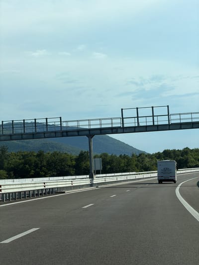 The image shows a highway with a white motorhome driving on it. The highway has a metal guardrail on the side and a large metal structure overhead, possibly for road signs or lighting. In the background, there are green hills covered in trees and a blue sky with some clouds. The scene appears to be in a rural area, and while the image is presented as being in the city of Peshterna, Bulgaria, there are no specific landmarks or details within the image to confirm this location.